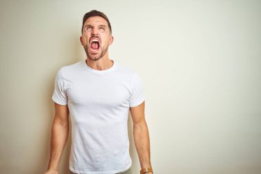 Genç yakışıklı adam izole arka plan kızgın ve deli çığlık sinirli ve öfkeli üzerinde rahat beyaz t-shirt giyen, öfke ile bağırarak. Öfke ve agresif kavram.