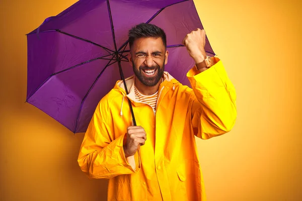 Young indian man wearing raincoat and purple umbrella over isolated yellow background annoyed and frustrated shouting with anger, crazy and yelling with raised hand, anger concept