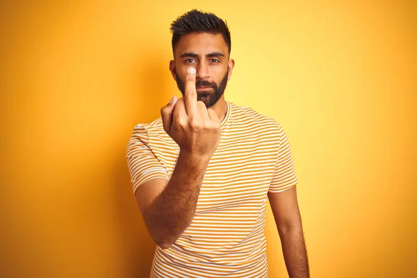 Young indian man wearing t-shirt standing over isolated yellow background Showing middle finger, impolite and rude fuck off expression