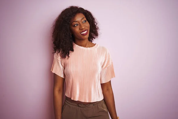 Young african american woman wearing t-shirt standing over isolated pink background looking away to side with smile on face, natural expression. Laughing confident.