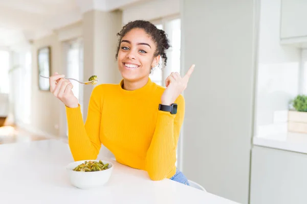 Young african american woman eating healthy green vegatables very happy pointing with hand and finger to the side