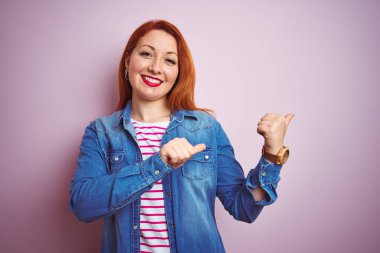 Denim gömlek giyen güzel kızıl saçlı kadın ve izole pembe arka plan üzerinde çizgili t-shirt el ve başparmak yukarı arka işaret, kendinden emin gülümseyerek