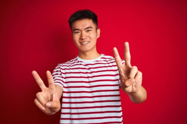 İzole kırmızı arka plan üzerinde duran çizgili t-shirt giyen genç Asyalı Çinli adam zafer işareti yapan parmakları gösteren kameraya bakarak gülümseyerek. İki numara.