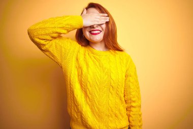 Güzel kızıl saçlı kadın izole sarı arka plan üzerinde duran kış kazak giyen gülümseyerek ve sürpriz için gözleri kaplayan yüzünde el ile gülüyor. Kör kavramı.