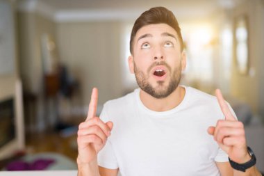 Evde rahat beyaz t-shirt giyen genç yakışıklı adam hayret ve yukarıya bakıyor ve parmakları ile işaret ve silah kaldırdı şaşırttı.