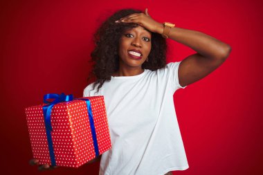 İzole kırmızı arka plan üzerinde duran doğum günü hediyesi tutan genç Afrikalı-Amerikalı kadın, utanç ve sürpriz yüz, kızgın ve sinirli ile şok el ile vurguladı. Korku ve hata için üzgün.
