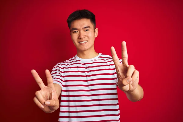 孤立した赤い背景の上に立つストライプのTシャツを着たアジアの若い中国人男性は 勝利のサインをしている指を示すカメラに微笑みかけている ナンバー2 — ストック写真