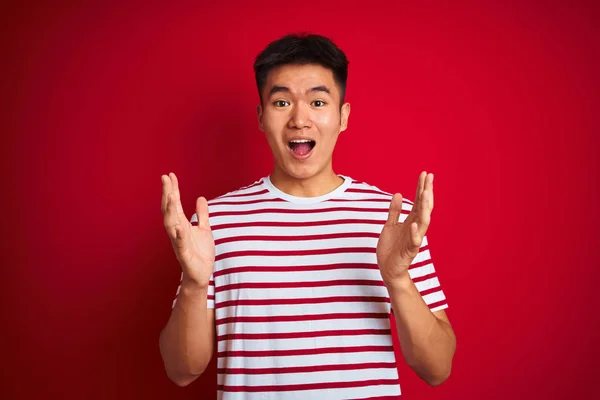 年轻的亚洲中国男子穿着条纹T恤站在孤立的红色背景庆祝疯狂和惊讶的成功与手臂抬起和睁开眼睛尖叫兴奋 优胜者概念 — 图库照片