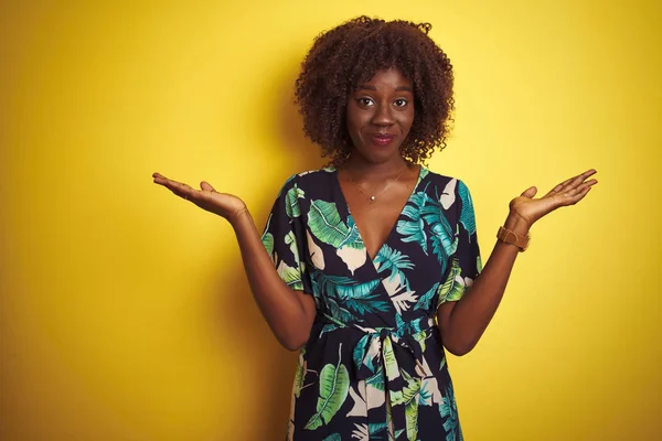 Young african afro woman wearing summer floral dress over isolated yellow background clueless and confused expression with arms and hands raised. Doubt concept.