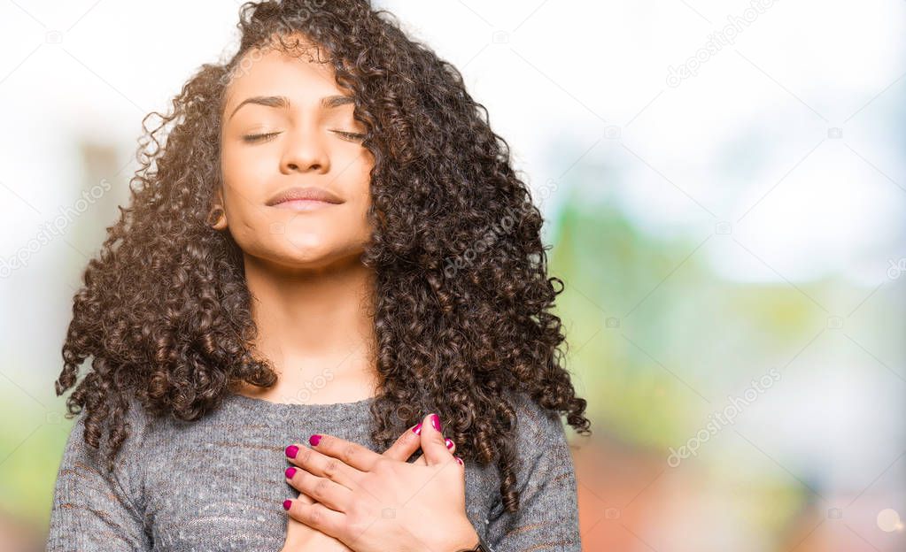 Young beautiful woman with curly hair wearing grey sweater smiling with hands on chest with closed eyes and grateful gesture on face. Health concept.