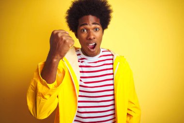 Afro saçlı afro saçlı bir adam izole sarı arka plan üzerinde duran yağmurluk giyen kızgın ve öfkeli öfke ile bağırırken sinirli ve öfkeli yumruğa yükselterek. Öfke ve agresif kavram.