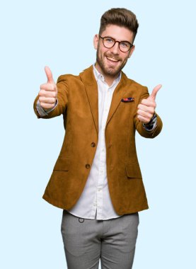 Onaylama gülümseyerek el, başparmak ile pozitif jest yapıyor ve başarı için mutlu gözlüklü genç yakışıklı iş adamı. Kamera, kazanan jest arıyorsunuz.