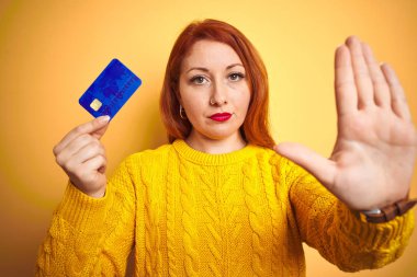 Genç müşteri redhead kadın ciddi ve kendinden emin ifade, savunma hareketi ile stop işareti yapıyor açık el ile sarı izole arka plan üzerinde kredi kartı tutan