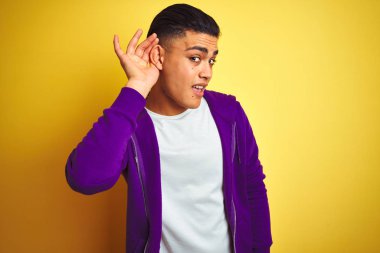 Mor sweatshirt giyen genç Brezilyalı adam izole sarı arka plan üzerinde ayakta kulak üzerinde el ile gülümseyerek söylenti veya dedikodu bir işitme dinlerken. Sağırlık kavramı.