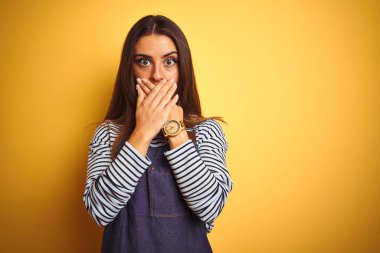 Izole sarı arka plan üzerinde duran önlük giyen genç güzel fırıncı kadın hata için elleri ile ağız kapsayan şok. Gizli kavram.