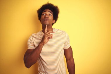 Afro saçlı amerikalı adam çizgili t-shirt izole sarı arka plan üzerinde duran giyen çene parmak ile şüphe hakkında konsantre ve merak bakarak