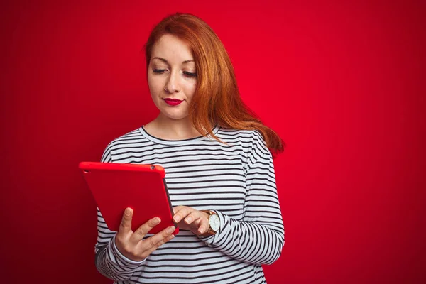 Young beautiful redhead woman using tablet over red isolated background with a confident expression on smart face thinking serious