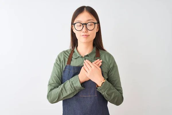 エプロンとメガネをかけた中国の若い店主女性は 目を閉じて感謝のジェスチャーをして胸に手を当て 孤立した白い背景の上に微笑む 健康の概念 — ストック写真