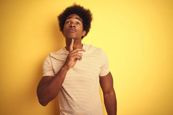 American man with afro hair wearing striped t-shirt standing over isolated yellow background Thinking concentrated about doubt with finger on chin and looking up wondering