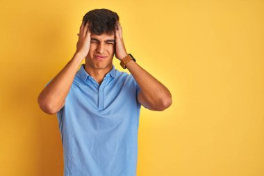 Mavi polo giyen genç hintli adam izole sarı arka plan üzerinde ayakta baş ağrısı umutsuz muzdarip ve stresli çünkü ağrı ve migren. Eller baş üstünde.