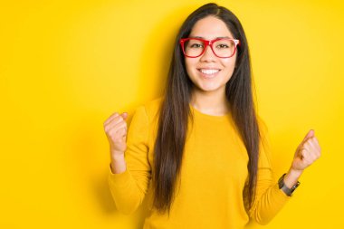 Güzel esmer kadın sarı izole arka plan üzerinde kırmızı gözlük giyen sürpriz ve kollarını kaldırdı ve açık gözleri ile başarı için şaşırttı kutluyor. Kazanan kavramı.