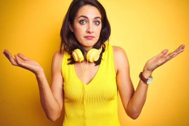 Genç güzel kadın sarı izole arka plan üzerinde kulaklık giyen habersiz ve kolları ve elleri kaldırdı ile karışık ifade. Şüphe kavramı.