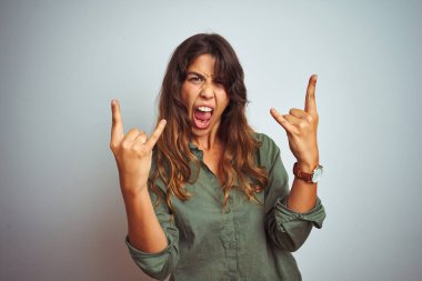 Gri izole arka plan üzerinde duran yeşil gömlek giyen genç güzel kadın elleri yukarı ile rock sembolü yapıyor deli ifade ile bağırarak. Müzik yıldızı. Ağır müzik konsepti.