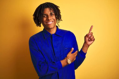 Yüzünde büyük bir gülümseme ile izole sarı arka plan üzerinde mekanik üniforma giyen dreadlocks ile Afro işçi adam, kameraya bakarak yan el ve parmak ile işaret.