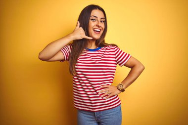 Genç güzel kadın çizgili t-shirt izole sarı arka plan üzerinde duran giyen telefonda konuşurken gibi el ve parmakları ile telefon jest yapıyor gülümseyerek. İletişim kavramları.