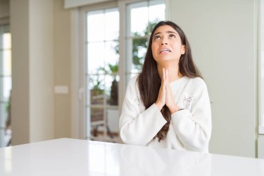 Genç güzel kadın evde beyaz masada dileniyor ve elleri ile birlikte dua ediyor umut la birlikte yüzünde umut ifade çok duygusal ve endişeli. Af dilemek istiyorum. Din kavramı.