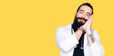 Uzun saçlı doktor tıbbi ceket ve stetoskop giyen yorgun rüya ve kapalı gözlerle gülümseyerek birlikte elleri ile poz.