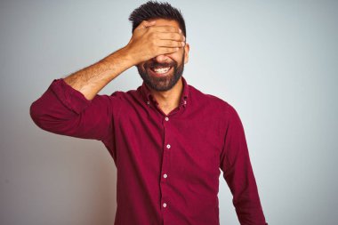 Kırmızı zarif gömlek giyen genç hintli adam izole gri arka plan üzerinde duran gülümseyerek ve sürpriz için gözleri kaplayan yüzünde el ile gülüyor. Kör kavramı.