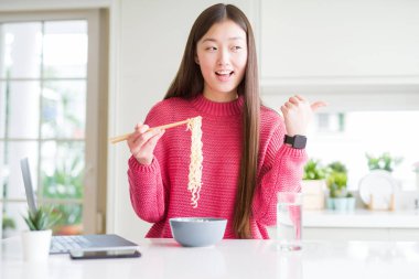 Güzel Asyalı kadın dizüstü bilgisayar kullanarak çalışan ve asya erişte yeme işaret ve mutlu yüzü gülümseyerek yan başparmak ile gösteren