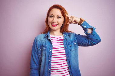 Denim gömlek giyen güzel kızıl saçlı kadın ve izole pembe arka plan üzerinde çizgili t-shirt gülümseyerek bir parmak, harika bir fikir ya da düşünce, iyi bellek ile baş işaret