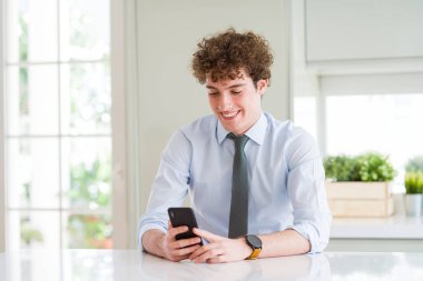 Ofiste akıllı telefonu kullanan genç iş adamı mutlu bir yüzle ayakta ve gülümseyerek dişlerini gösteren kendinden emin bir gülümsemeyle