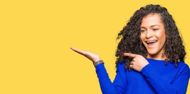 Kıvırcık saçlı giyen kış kazak hayret ve el ile sunulması ve parmak ile işaret ederken kameraya gülümseyen genç güzel kadın.