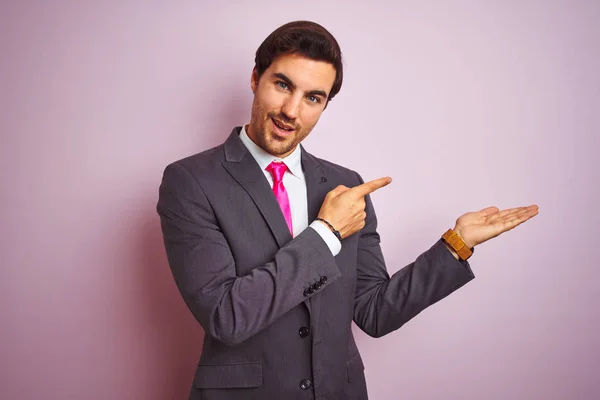 年轻的英俊商人穿着西装和领带站在孤立的粉红色背景惊讶和微笑的相机 同时呈现与手和手指指点 — 图库照片