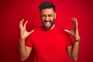 Genç yakışıklı hintli adam izole kırmızı arka plan üzerinde t-shirt giyen kollarını kaldırdı ve kapalı gözleri heyecan çığlık ile başarı için deli ve deli kutluyor. Kazanan kavramı
