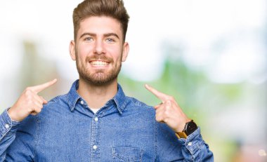 Kendinden emin gösterilen ve işaret parmakları diş ve ağız ile gülümseyen rahat kot gömlek giymiş genç yakışıklı sarışın adam. Sağlık kavramı.