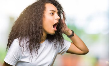 Genç güzel kız bağırarak ve ağzı üzerinde el ile yan için çığlık casual beyaz t-shirt giyen kıvırcık saçlı. İletişim kavramı.