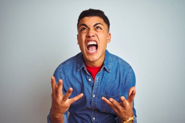Denim gömlek giyen genç brezilyalı adam izole beyaz arka plan üzerinde duran deli ve deli bağırarak ve agresif ifade ve kollar kaldırdı ile bağırıyor. Hayal kırıklığı kavramı.