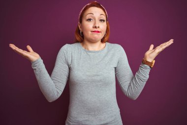 Güzel kızıl saçlı kadın gri t-shirt giyen ve izole mor arka plan üzerinde diadem habersiz ve kollar ve eller ile karışık ifade kaldırdı. Şüphe kavramı.