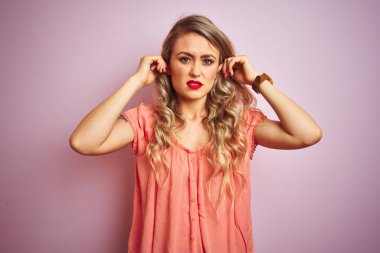 Genç güzel kadın yüksek sesle müzik sesi için rahatsız ifade ile parmakları ile kulakları kapsayan pembe izole arka plan üzerinde duran t-shirt giyiyor. Sağır kavramı.