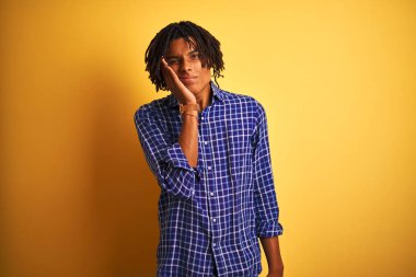 Izole sarı arka plan üzerinde duran rahat gömlek giyen dreadlocks ile Afro adam yorgun görünümlü ve çapraz kolları ile depresyon sorunları ile sıkılmış düşünme.