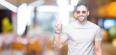 Genç yakışıklı adam mutlu yüz zafer işareti yaparken kameraya göz kırpıyor ile gülümseyen izole arka plan üzerinde güneş gözlüğü takıyor. İki numaralı.