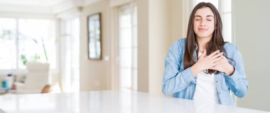 Göğüs Kapalı gözlerle ellerinde ve yüzünde minnettar jest ile geniş açı resim güzel genç kadının evde beyaz masaya oturan gülümsüyor. Sağlık kavramı.