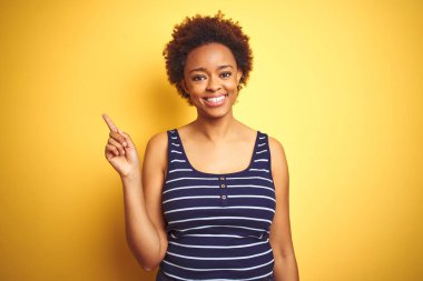 Beauitul afro-amerikan kadın yüzünde büyük bir gülümseme ile izole sarı arka plan üzerinde yaz t-shirt giyen, kameraya bakarak yan el parmağı ile işaret.