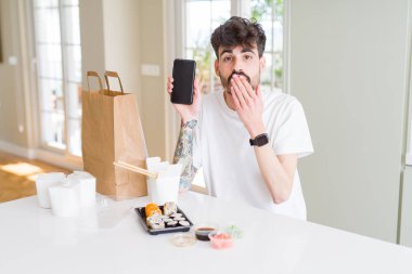 Genç adam eve teslim asya suşi yeme ve hata için utanç ile şok el ile akıllı telefon ekran kapağı ağız gösteren, korku ifadesi, sessizlik, gizli kavram korkmuş