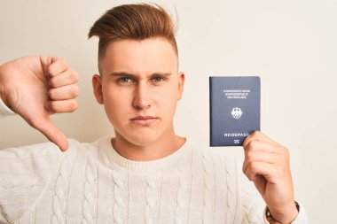 Kızgın yüz ile izole beyaz arka plan üzerinde Almanya Alman pasaportu tutan genç yakışıklı adam, başparmak aşağı, ret kavramı ile sevmediğimi gösteren negatif işareti
