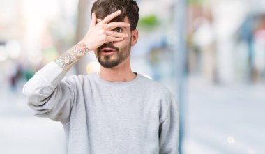 Mahcup ifadeyle parmaklarımın arasından seyir yüz ve gözler el ile kapsayan şok bakmak izole arka plan üzerinde sweatshirt giyen genç yakışıklı adam.